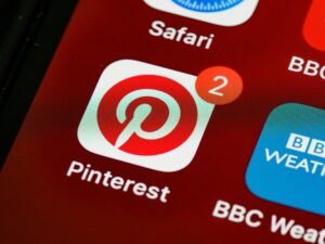 Макрофотографія екрана смартфона, що відображає іконку програми Pinterest (біле коло з червоною літерою 'P') з червоним значком повідомлення, що показує цифру '2'. Частково видно іконки Safari та BBC Weather.